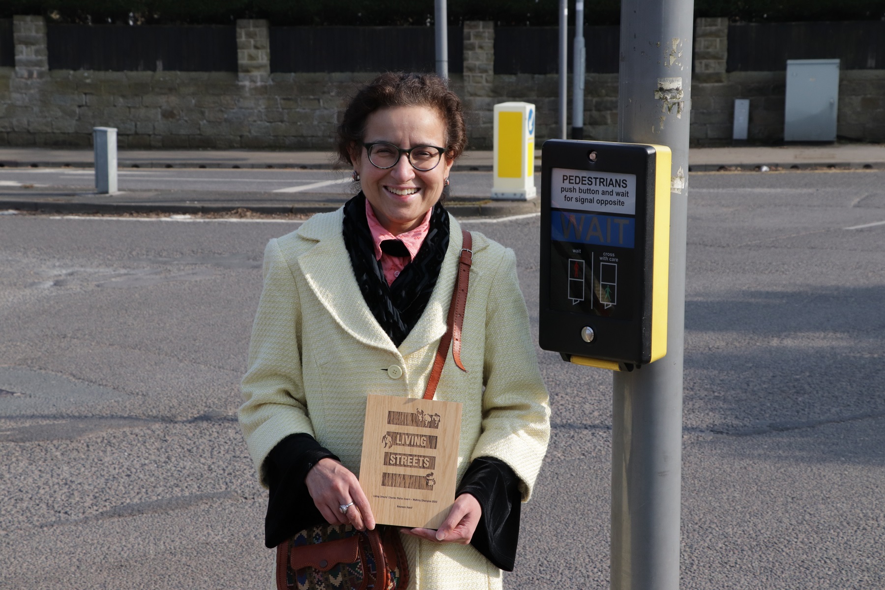 Nasreen Hanif is standing by a pedestrian crossing holding a wooden plaque