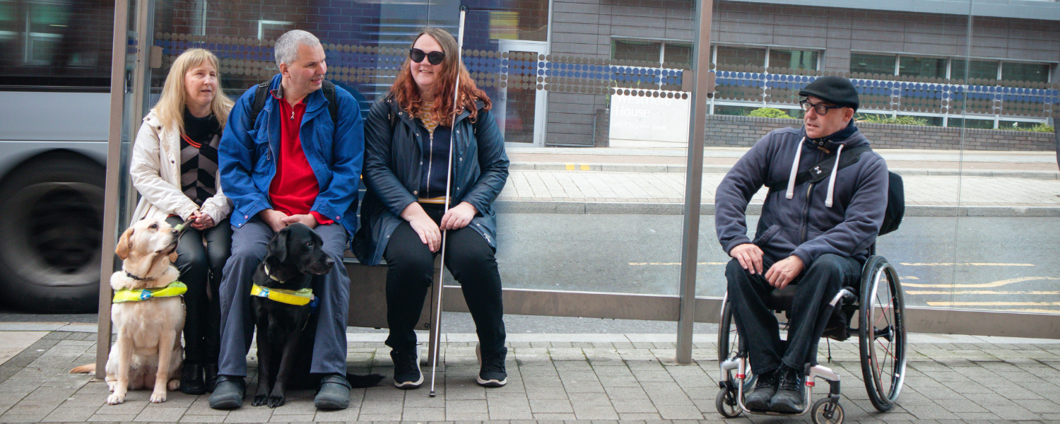 3 blind people sit at a bus stop, one has a guide dog, there is also a man sitting in a wheelchair