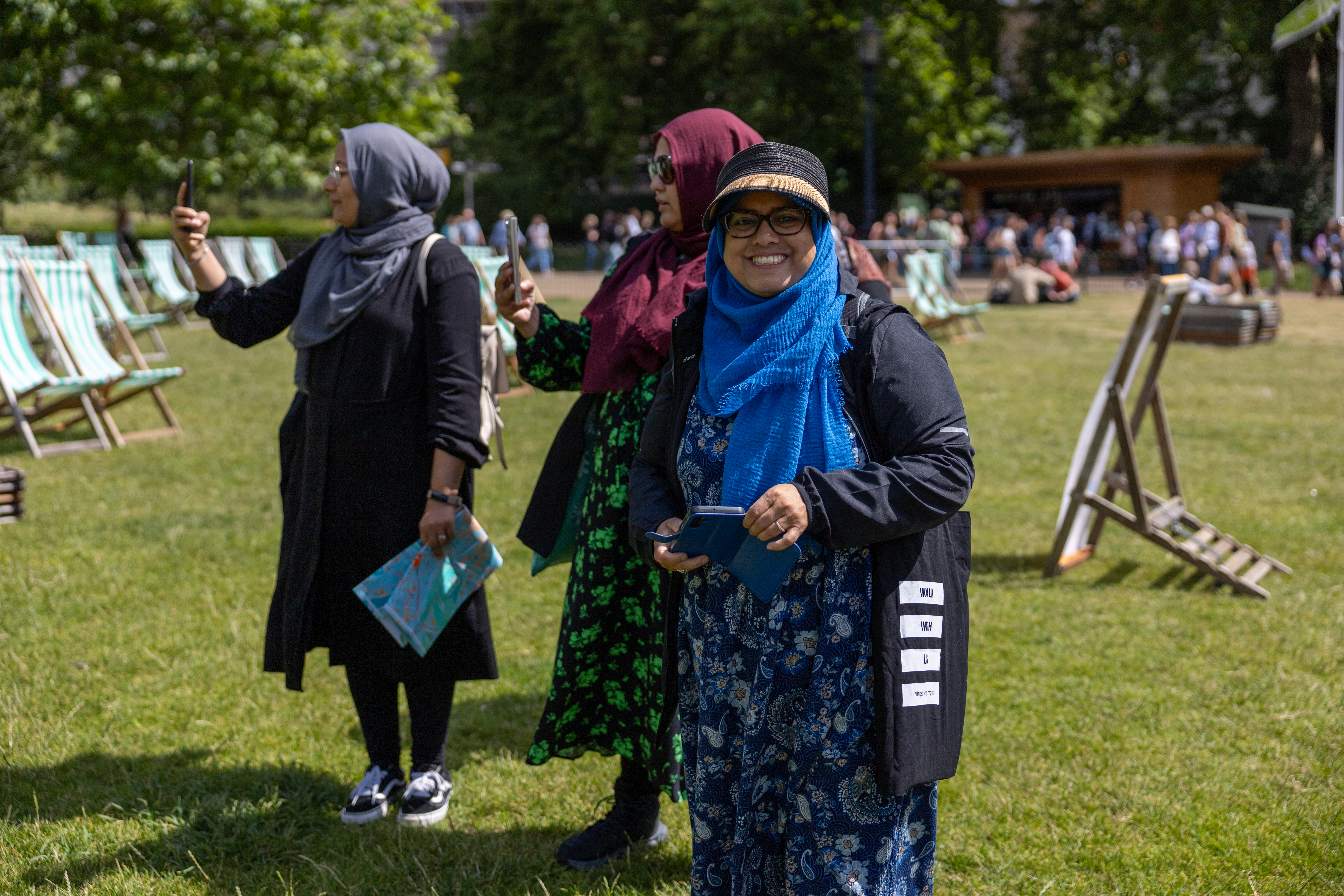 Three women dressed in hijabs stand in a park, all three have phones in their hand and one is smiling directly at the camera