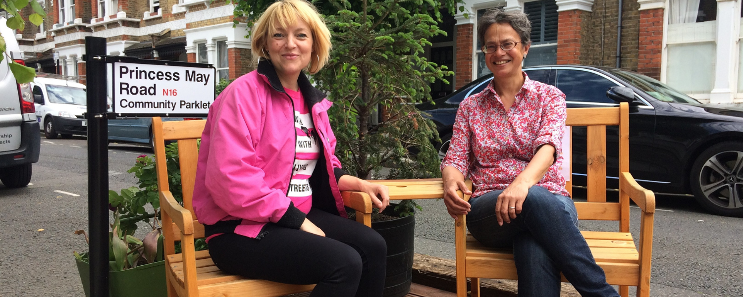two women sit on a bench in a parklet, one is a Living streets staff member