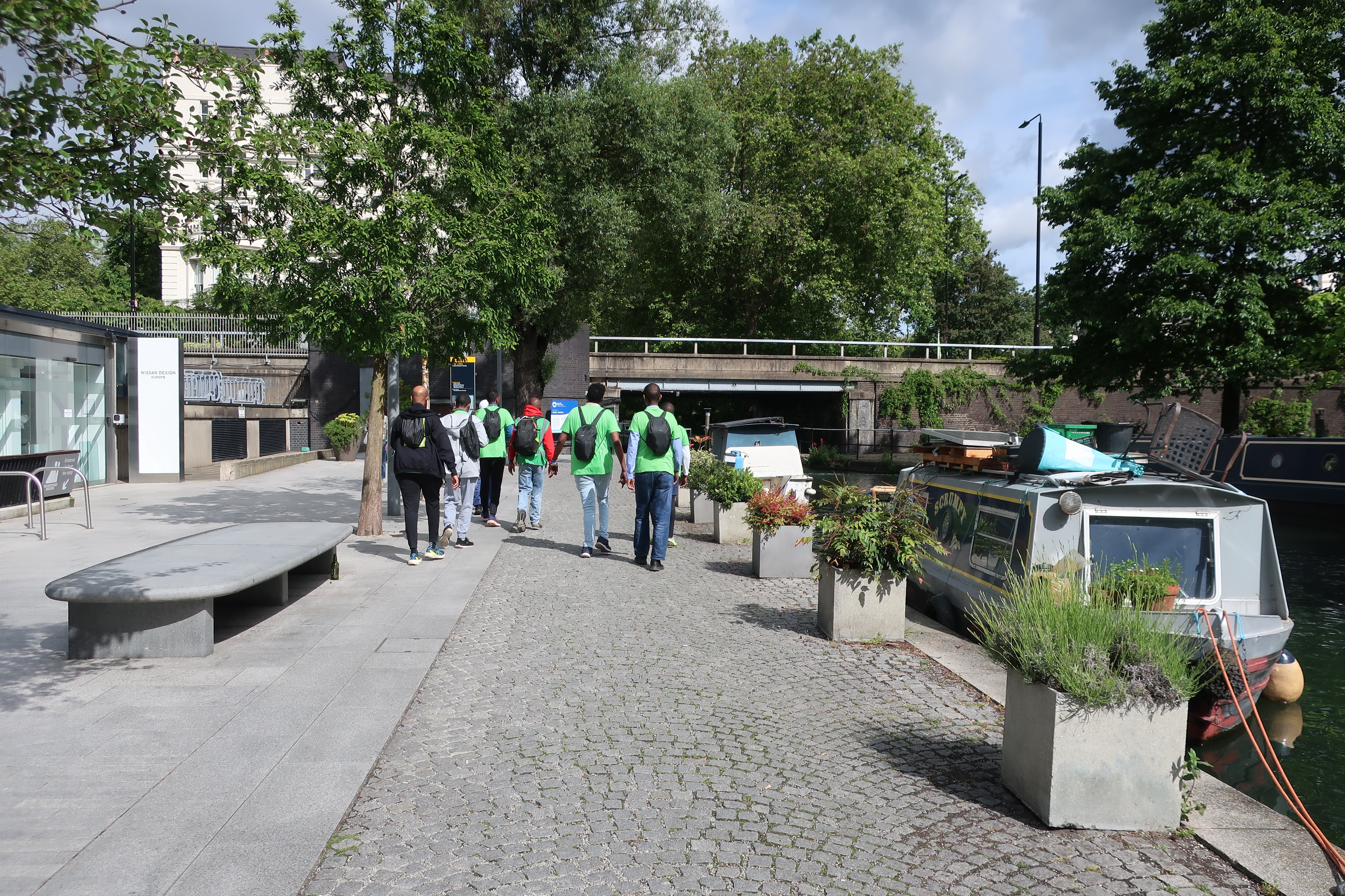 a group of men in living streets tshirts walk by a canal path on a sunny day