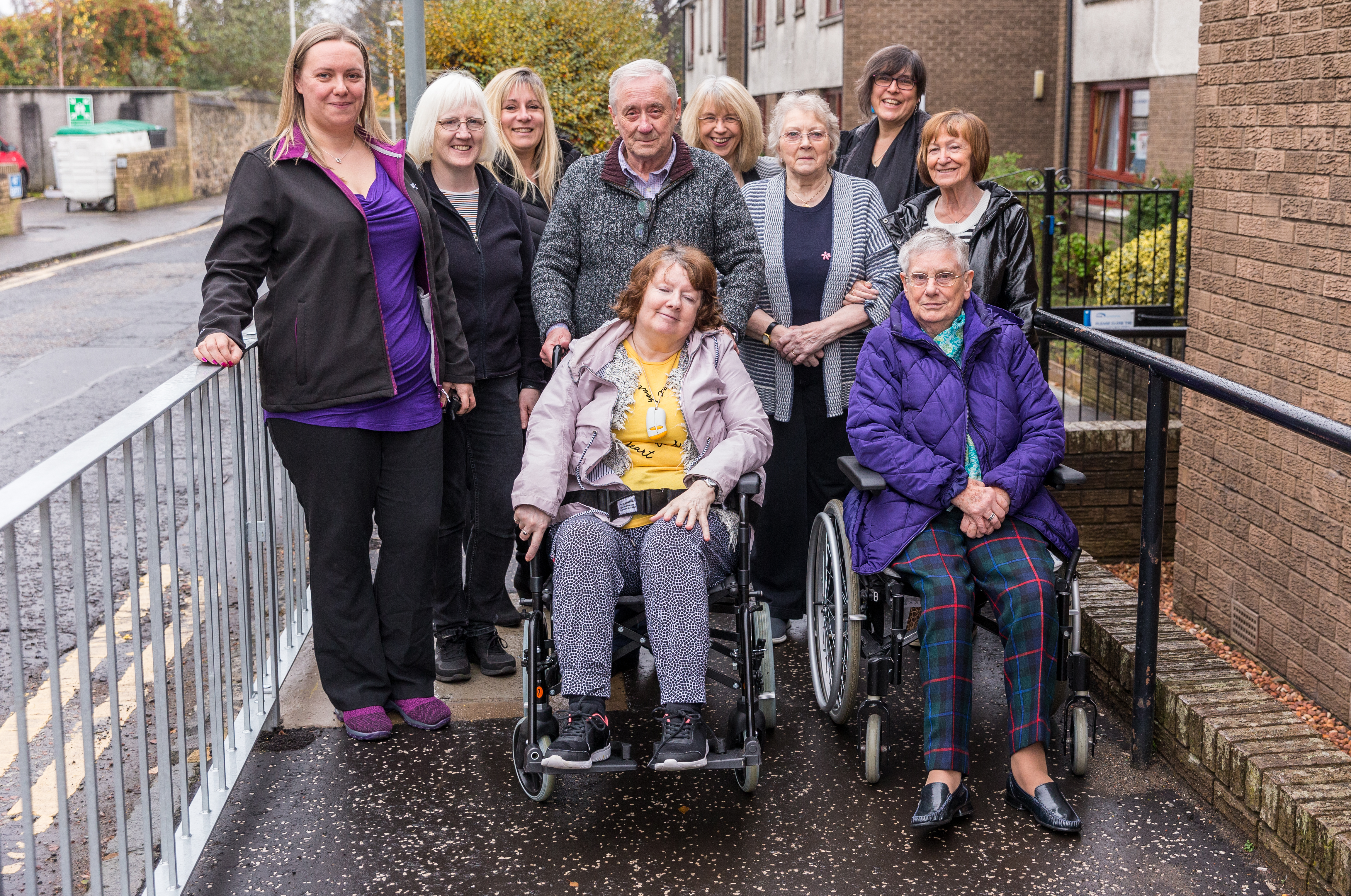 a group of older people including two women in wheelchairs, stand and smile at the camera on the pavement