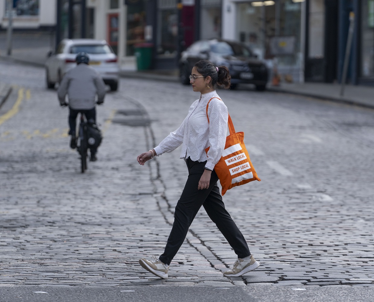 An image of a person walking across a road. They are holding a tote bag which reads Walk Local, Shop Local