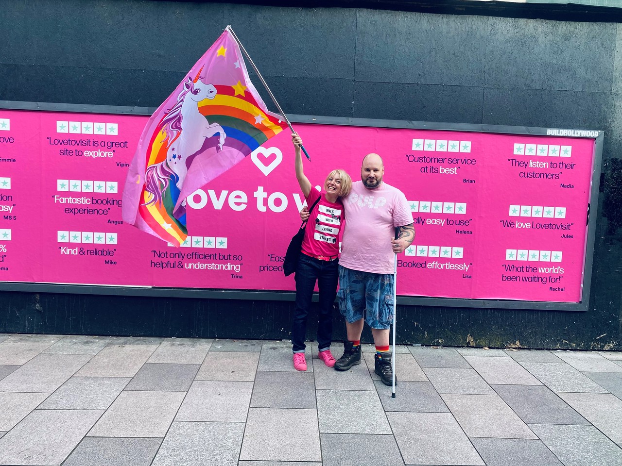 Aisha and Justin stand infront of a pink sign and Aisha is holding a unicorn flag