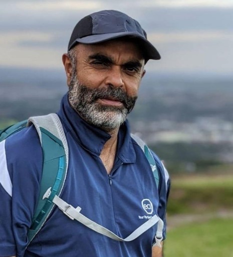 Mustafa Desai is standing with mountains behind him wearing a blue hat, top and bag