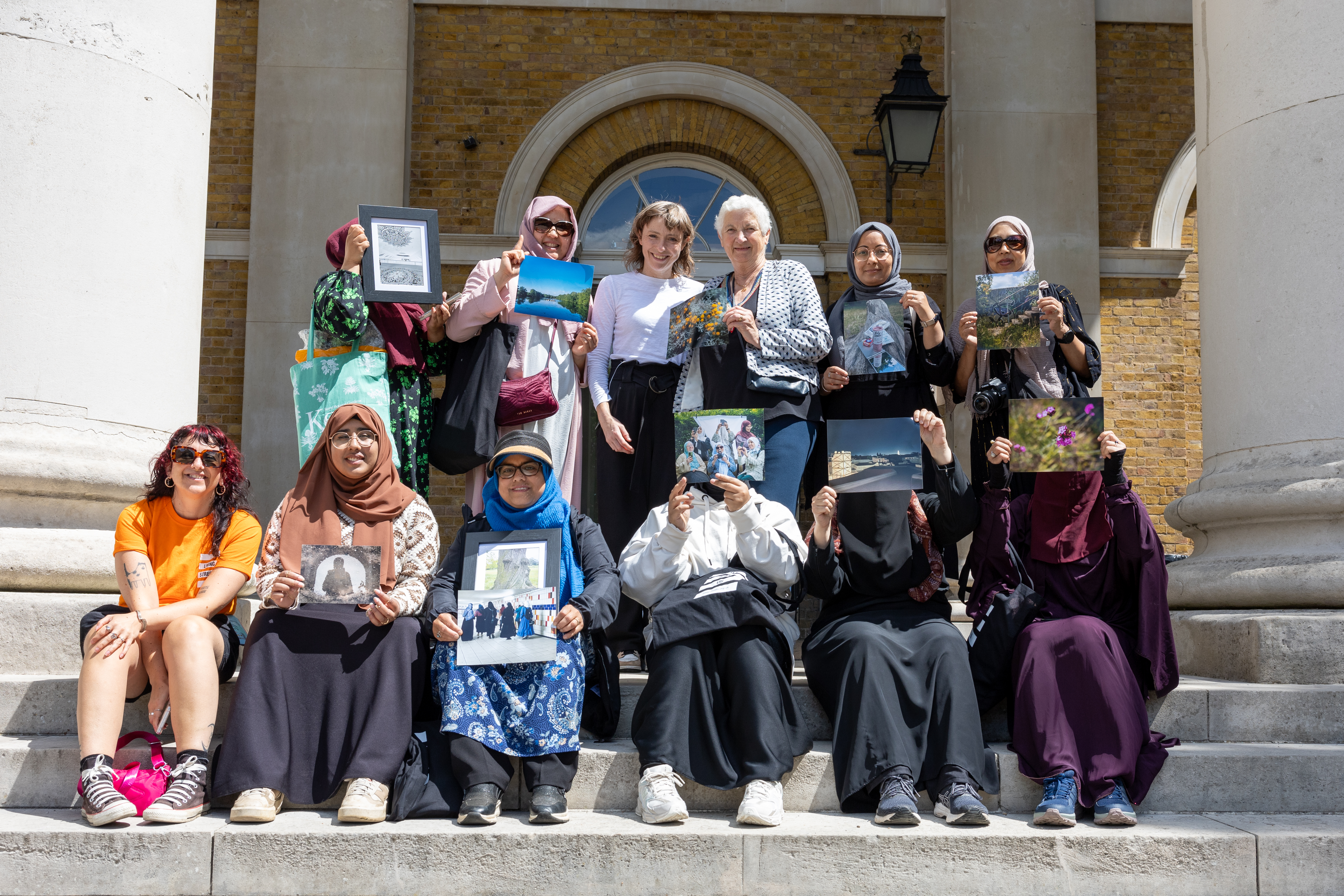 A group of women are outside an art gallery, the back row are standing up, the front sitting down. Some are holding art