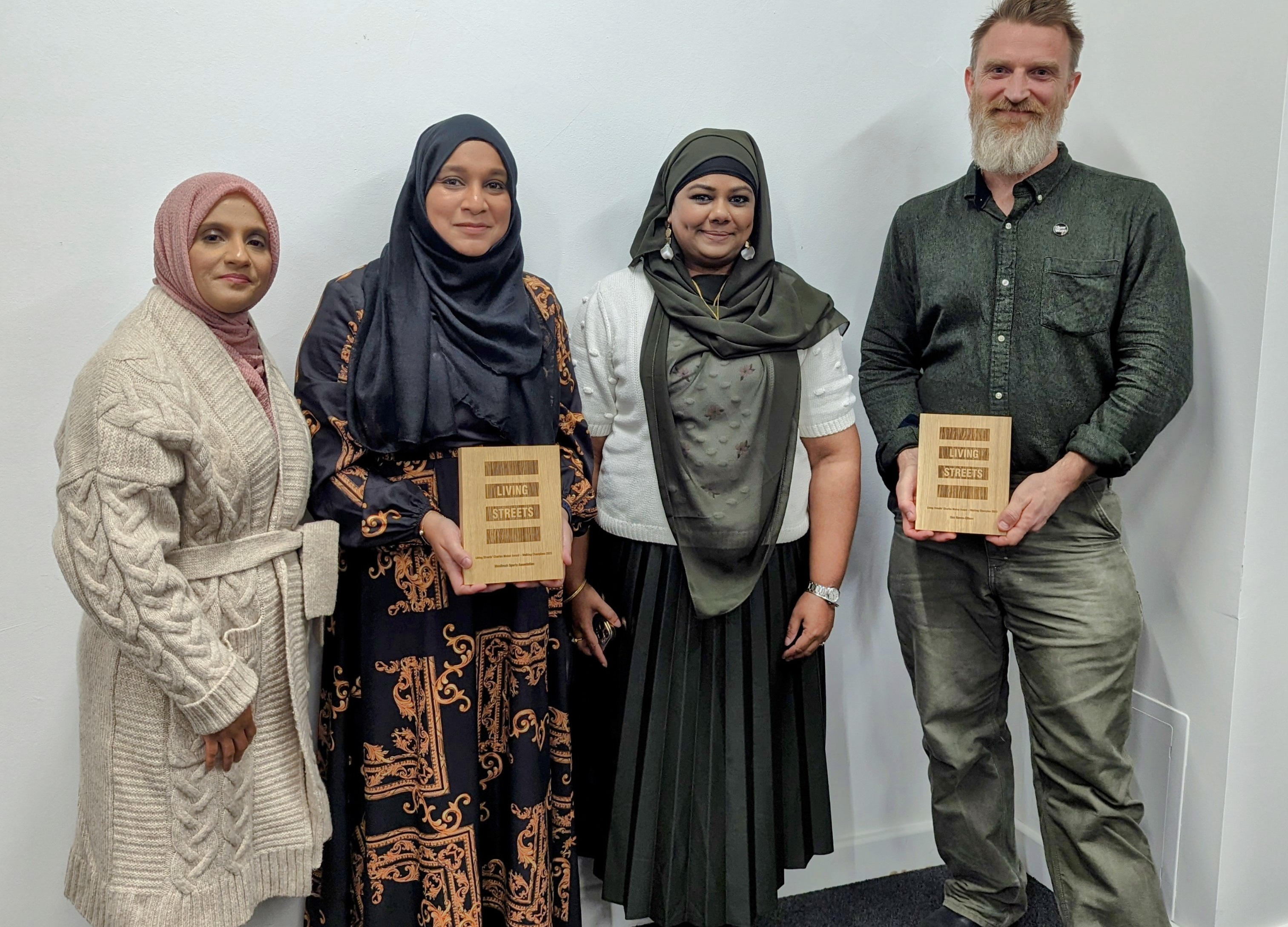 three women in hijabs are standing together, the woman in the middle is holding a wooden plaque. A man is standing on the right of the woman and is also holding a wooden plaque.