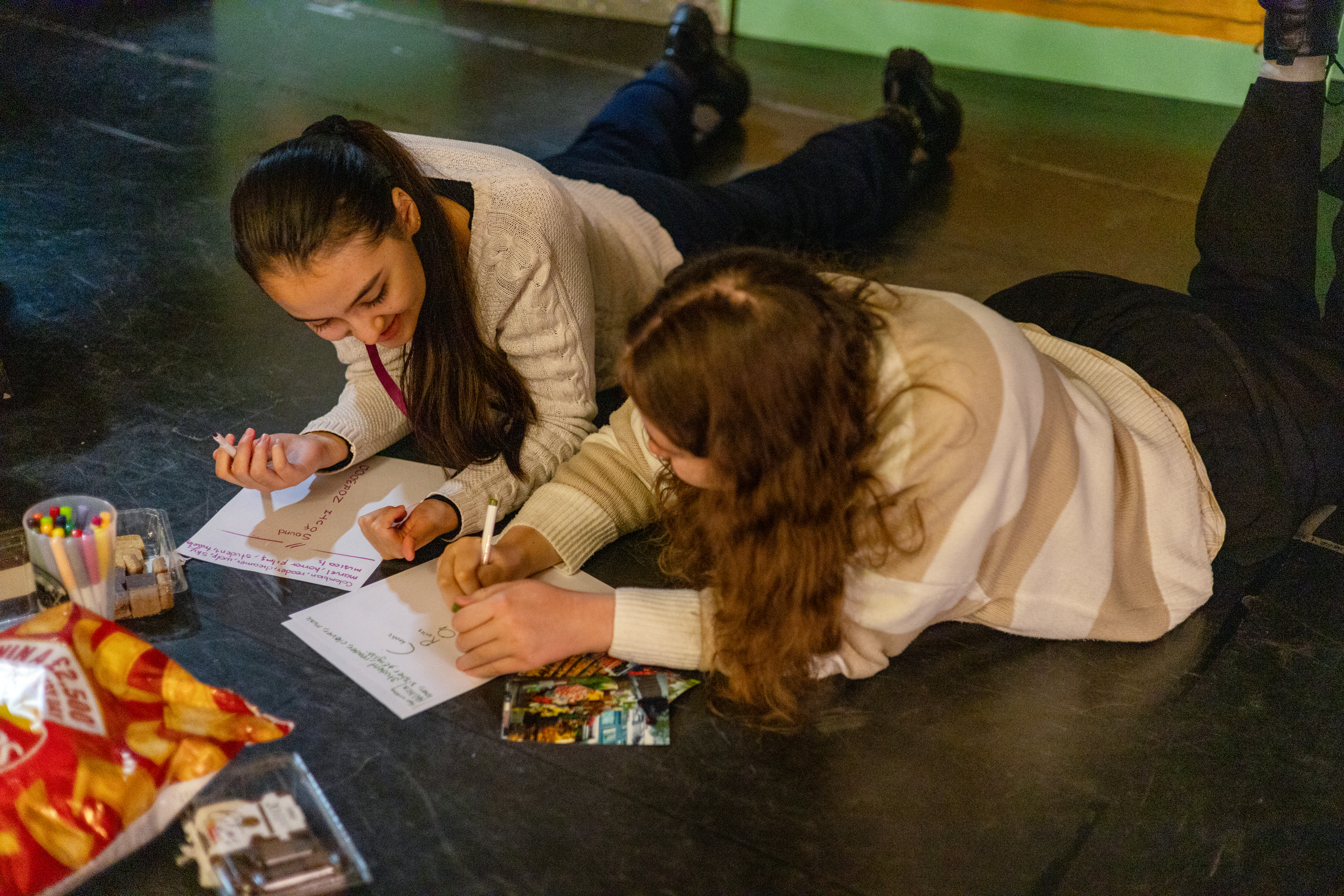 two girls lie on the floor and work together using pens and paper