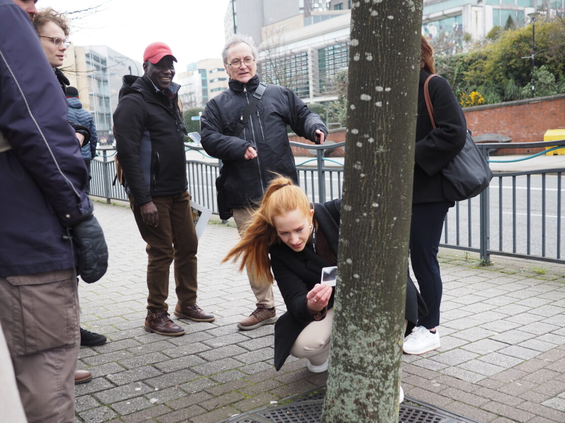a woman bends to observe lichen on a tree surrounded by a group of people